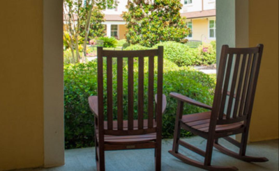 The Beacon at Gulf Breeze, a view of two wood rocking chairs overlooking green bushes, trees and grass outside.