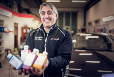 A Color Glo employee holds up colored bottles with a car and shop behind him.