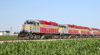 A train of The Iowa Northern Railway Company travels across green fields with farm silos in the background.