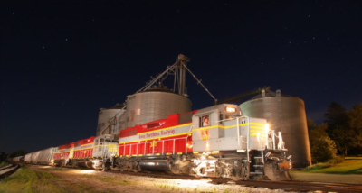 An Iowa Northern Railway Company train traveling at night with lights on and metal cylinder building behind it.