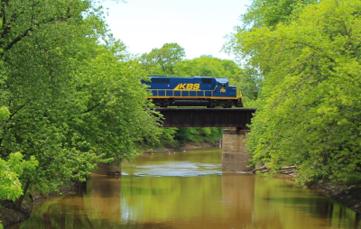 A Kankakee, Beaverville & Southern Railroad train going over a river on a small bridge with trees lining both sides of the river.