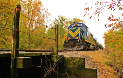 A Kankakee, Beaverville & Southern Railroad train about to pass over a small bridge with wood posts and wire along the sides.