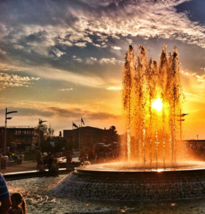 A beautiful view of a fountain with an orange sun and cloudy skies behind it in Owensboro Kentucky.
