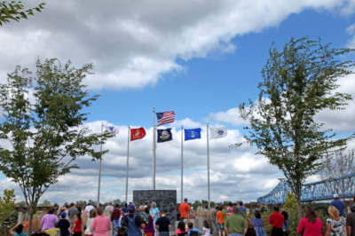 A crowd of people stand before 6 flag poles and an iron truss bridge in the left background.