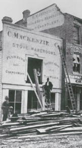 Mackenzie Milne. Old black and white photo showing people working at a Mackenzie building with piles of wood and ladders up.