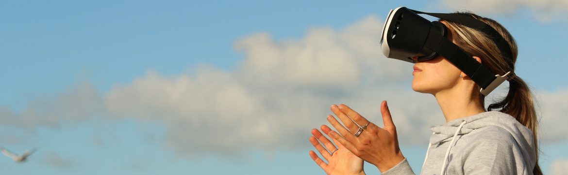 A woman wearing a virtual reality headset outside with her hands in an open gesture in front of her. Cloud and blue sky in the background.