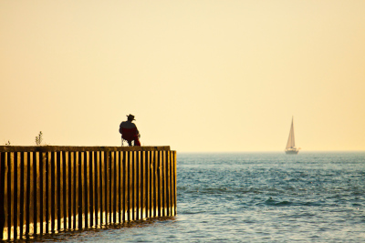 Goderich, Ontario; A min sitting on a pier with a sailboat in the distance on the water.