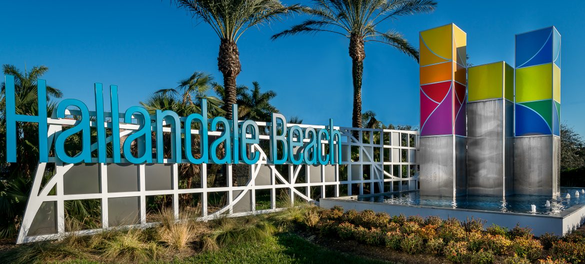 Hallandale Beach, Florida sign to the city with metallic art on the right and palm trees.