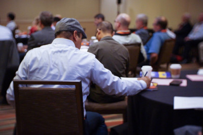 Aluminum Extruders Council. A man writing during a convention with other people at tables in the room all around.