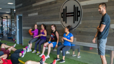 Core Progression. People leaning against a wall squatting and passing a ball while others sit on the floor exercising and a trainer watches.