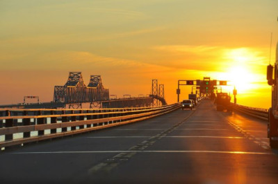 Sustainable Maryland. View down a bridge with a few cars in the distance and a bright sun on the right near the horizon.