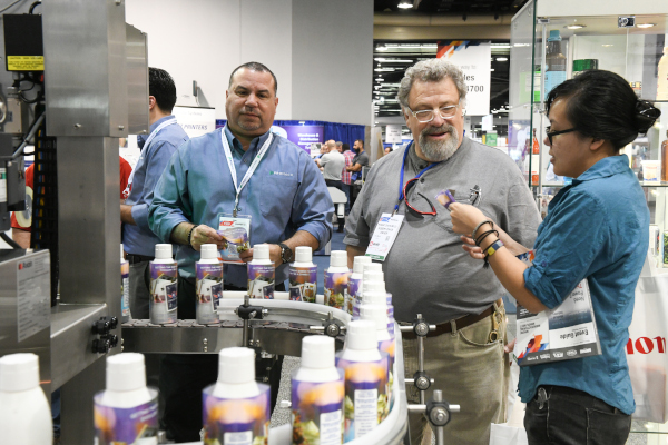 MD&M West Expo. 3 People at an event looking at a machine with cans of product in a semicircular row.