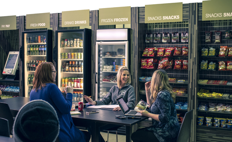 365 Retail Markets; 3 people sitting at a table with different shelves and refrigeration units along the wall behind with drinks and snacks.