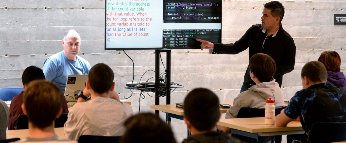 A group of young people in a room with an adult pointing at code on a screen in front of them.