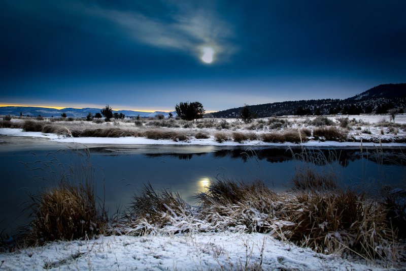 Klamath County, Oregon photo of a river with the sun behind a small wispy cloud, snow on the ground and hills in the distance.