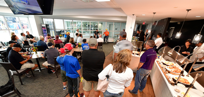 Richmond Raceway Complex event photo showing two men on one side of the room with a microphone and people sitting and standing with catering on the right.