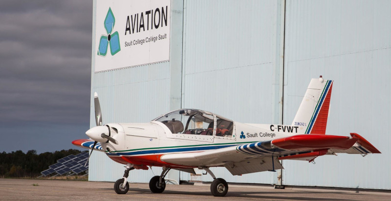 Sault Ste. Marie Airport hangar with a Sault College sign and airplane parked out front of it.