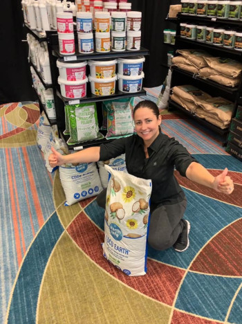 Greenstar Plant Products Inc. ; a woman in a store with a product bag on the ground, she is kneeling with her arms stretched open.