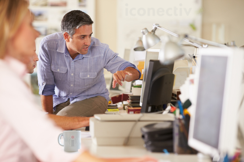 iconectiv a man in the background pointing at a monitor with a blurry woman in the foreground working on a computer.