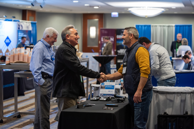 The Association for Manufacturing Excellence - AME conference showing two men shaking hands over a table and other people around the room.