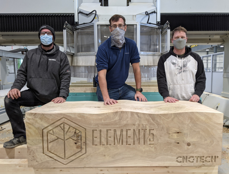 Element5 CNC area with 3 employees posing for a photos by a block of wood with Element5 logo etched into it.