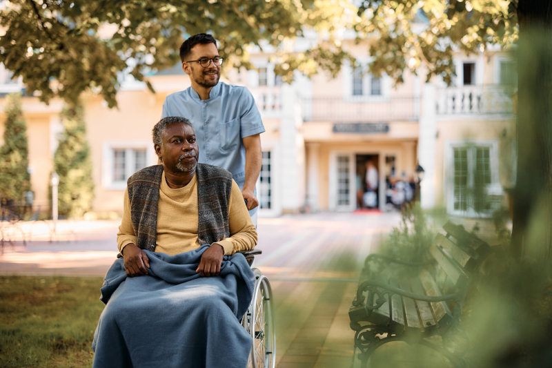 African American senior in wheelchair being pushed by male caregiver in the park of residential care home. Copy space.